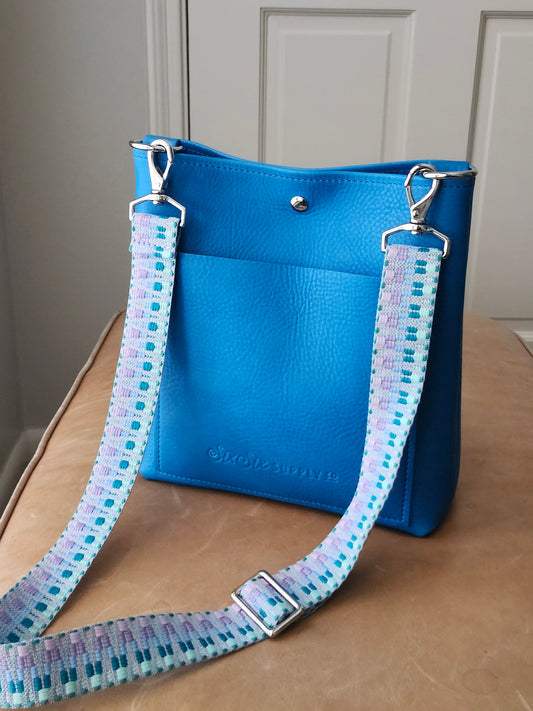 Blue crossbody handbag with a pastel blue patterned webbing strap on a leather surface